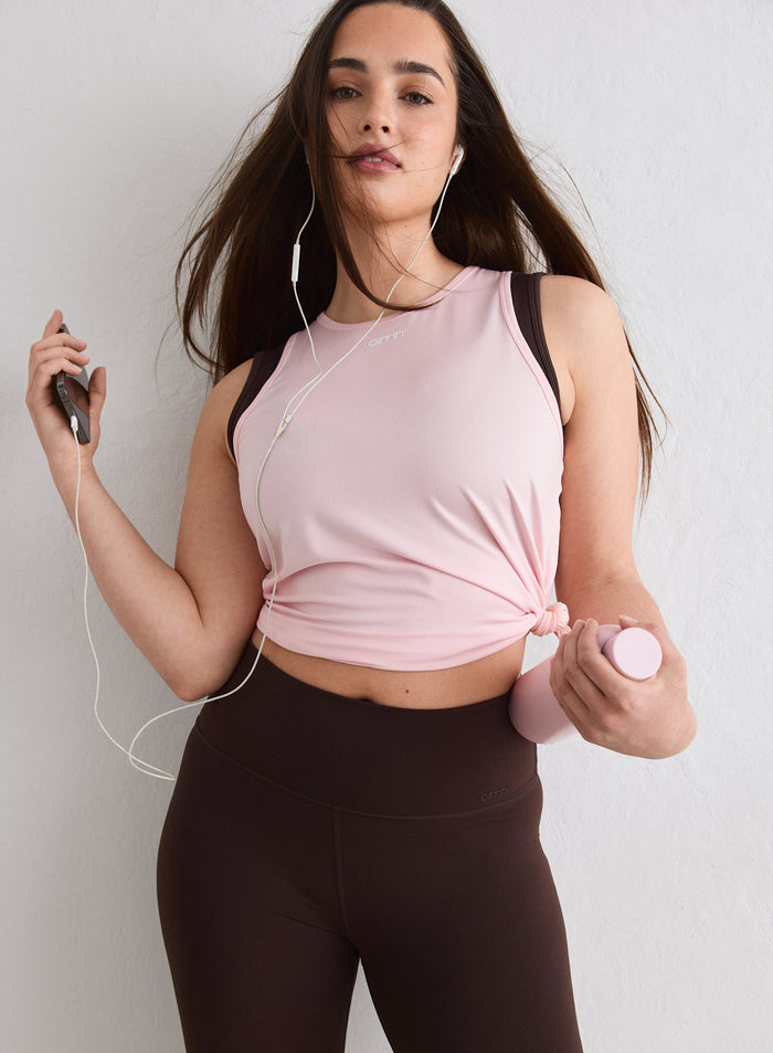 Nainen yllään vaaleanpunainen, vyötärölle solmittu aim'n Soft Basic Singlet, ruskeat leggingsit sekä puhelin ja vesipullo [L-174cm]