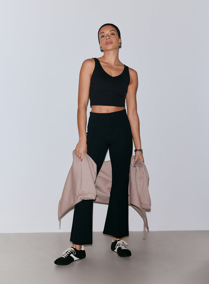 Nainen yllään mustat Luxe Kick Flare Leggings -leggingsit ja musta v-kaula-aukkoinen crop-toppi, pitäen vaaleanpunaista vetoketjullista takkia käsivarrellaan [36-170CM]