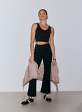 Nainen yllään mustat Luxe Kick Flare Leggings -leggingsit ja musta v-kaula-aukkoinen crop-toppi, pitäen vaaleanpunaista vetoketjullista takkia käsivarrellaan [36-170CM]