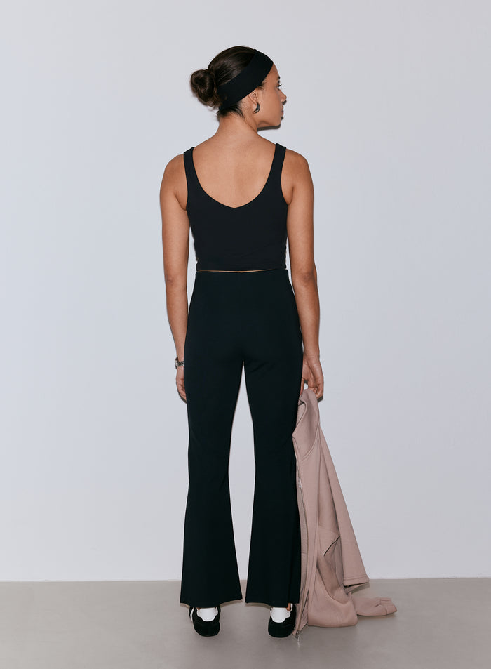 Nainen takaapäin, yllään mustat Luxe Kick Flare Leggings -trikoot ja musta crop top, pidellen vaaleanpunaista takkia. [36-170CM]