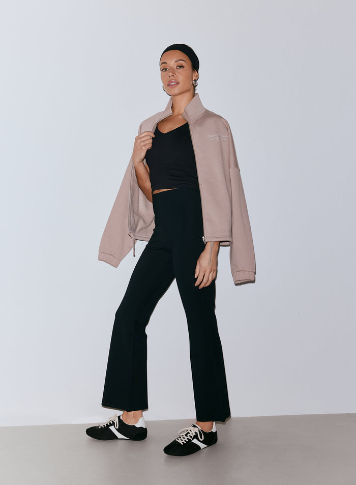 Nainen yllään mustat Luxe Kick Flare Leggings -leggingsit, musta crop-toppi ja vaaleanruskea vetoketjullinen takki olkapäillään, mustalla päänauhalla ja mustavalkoisilla tennareilla. [36-170cm]