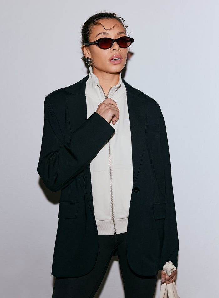 Nainen yllään musta Luxe Relaxed Blazer, vaalea vetoketjullinen toppi ja aurinkolasit [40-170CM]