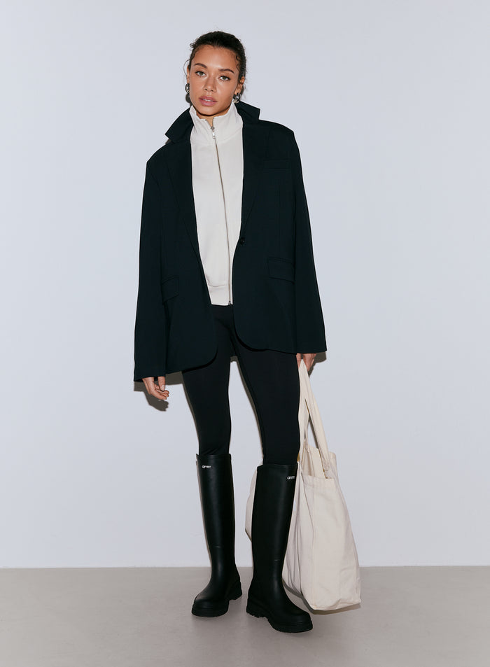 Nainen yllään musta Luxe Relaxed Blazer valkoisen vetoketjullisen topin päällä, mustat leggingsit ja mustat polvipituiset saappaat, pitäen kädessään kermanväristä kangaskassia [40-170CM]