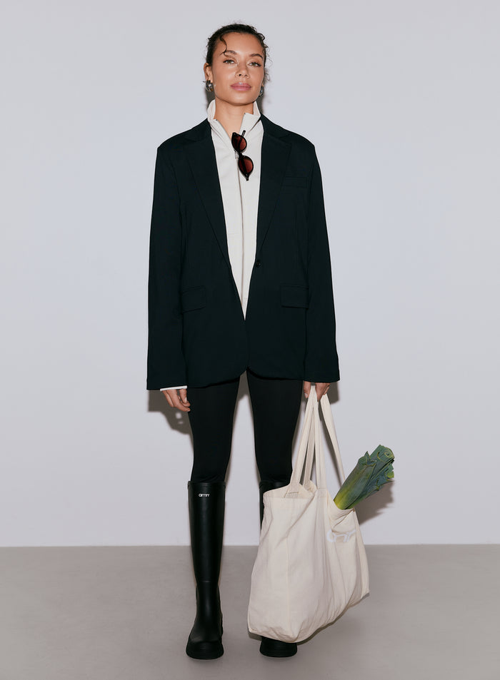 Nainen yllään musta Luxe Relaxed -bleiseri, valkoinen vetoketjullinen toppi, mustat leggingsit ja mustat saappaat, pidellen kangaskassia, jossa on purjo [40-170CM]