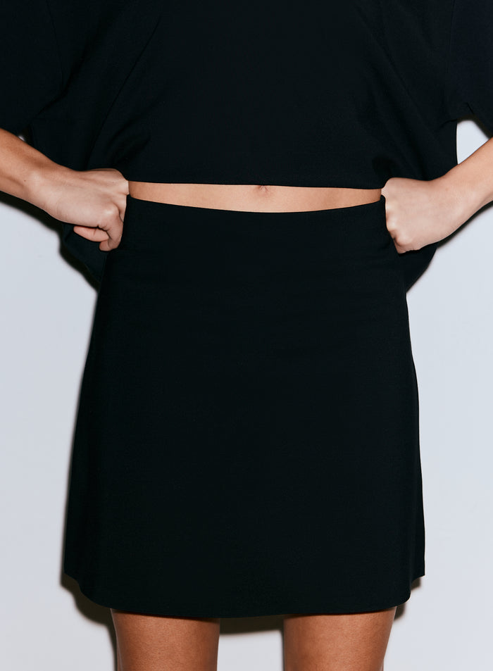 Lähikuva henkilöstä, jolla on yllään musta Luxe Mini Skort, vetäen vyötärönauhaa ylöspäin [36-170CM]