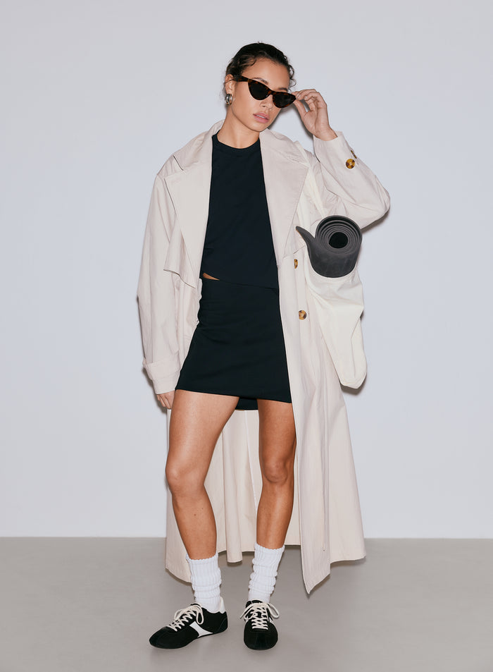 Nainen mustassa Luxe Mini Skortissa, mustassa topissa ja beigessä trenssitakissa joogamaton kanssa [36-170CM]