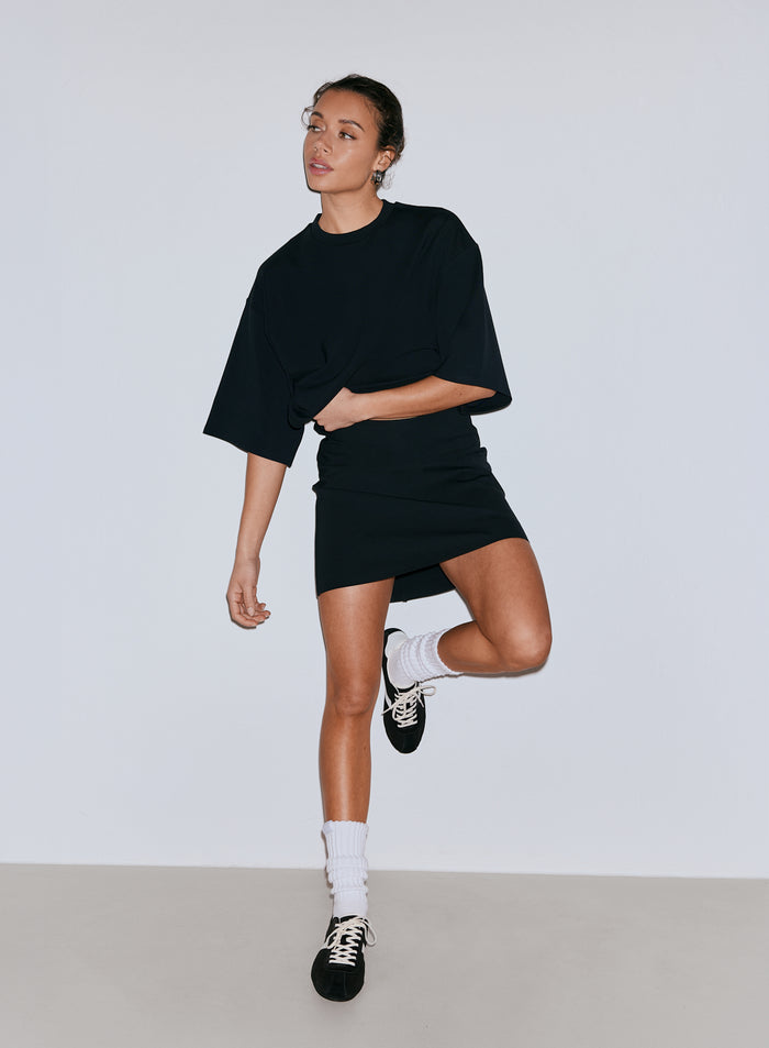 Malli pukeutunut mustaan oversized t-paitaan ja mustaan Luxe Mini Skortiin. [36-170CM]