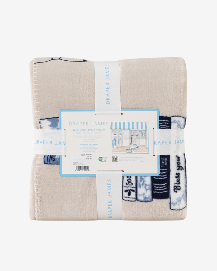 A folded beige decorative throw blanket with blue and white striped and knit patterns, wrapped in a white paper band labeled 'DRAPER JAMES' with its retail tag in the center, on a plain white background.