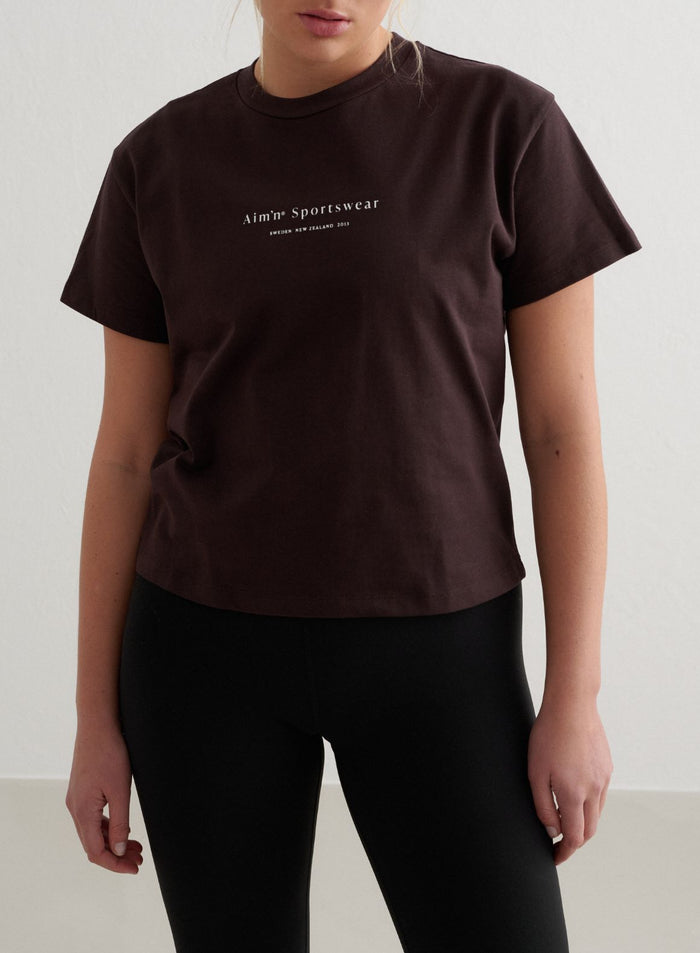 Tummanruskea puuvillainen Serif Boxy T-paita, jossa on valkoinen aim'n-logo ja pyöreä resoripääntie [S - 172 cm]