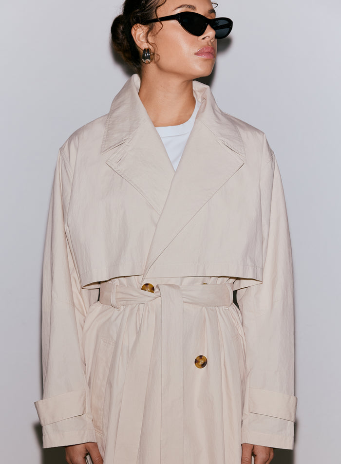 Naisesta lähikuva, yllään café au lait -värinen aim'n Femme Trenchcoat, jossa on sidottu vyö ja suuret napit, yhdistettynä mustiin aurinkolaseihin. [M-170cm]