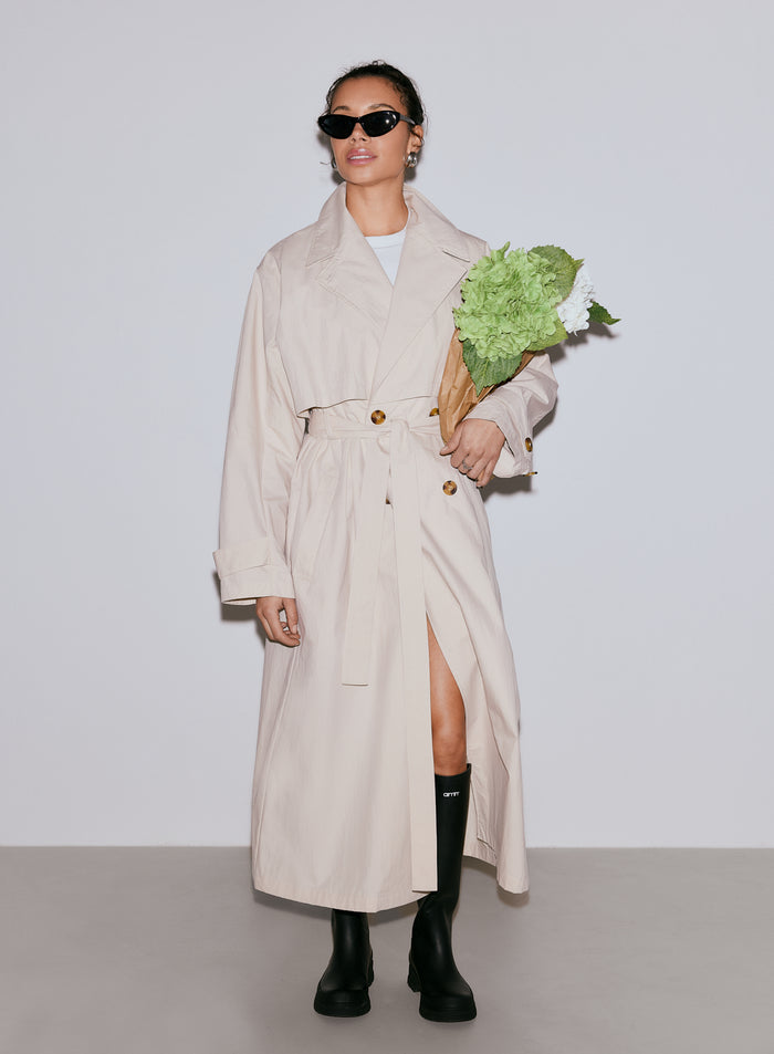 Nainen yllään pitkä café au lait -värinen Femme Trenchcoat -trenssitakki, mustat aurinkolasit ja mustat saappaat, kädessään kimppu vihreitä ja valkoisia kukkia [M-170cm]