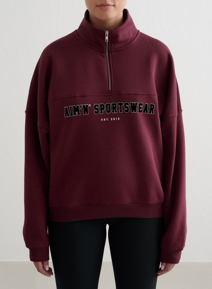 Kirsikanpunainen Varsity Sweat Half Zip pystykauluksella ja aim'n sportswear -kirjailulla.