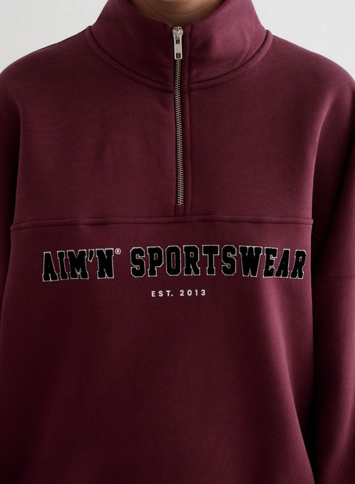 Kirsikanpunainen Varsity Sweat Half Zip pystykauluksella ja mustalla varsity-tyylisellä aim'n-rintabrodeerauksella.