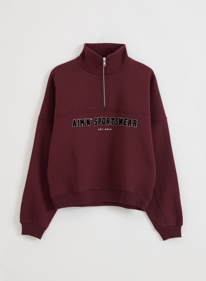 Kirsikanpunainen Varsity Sweat Half Zip, jossa on varsity-tyylinen aim'n-painatus ja pystykaulus.