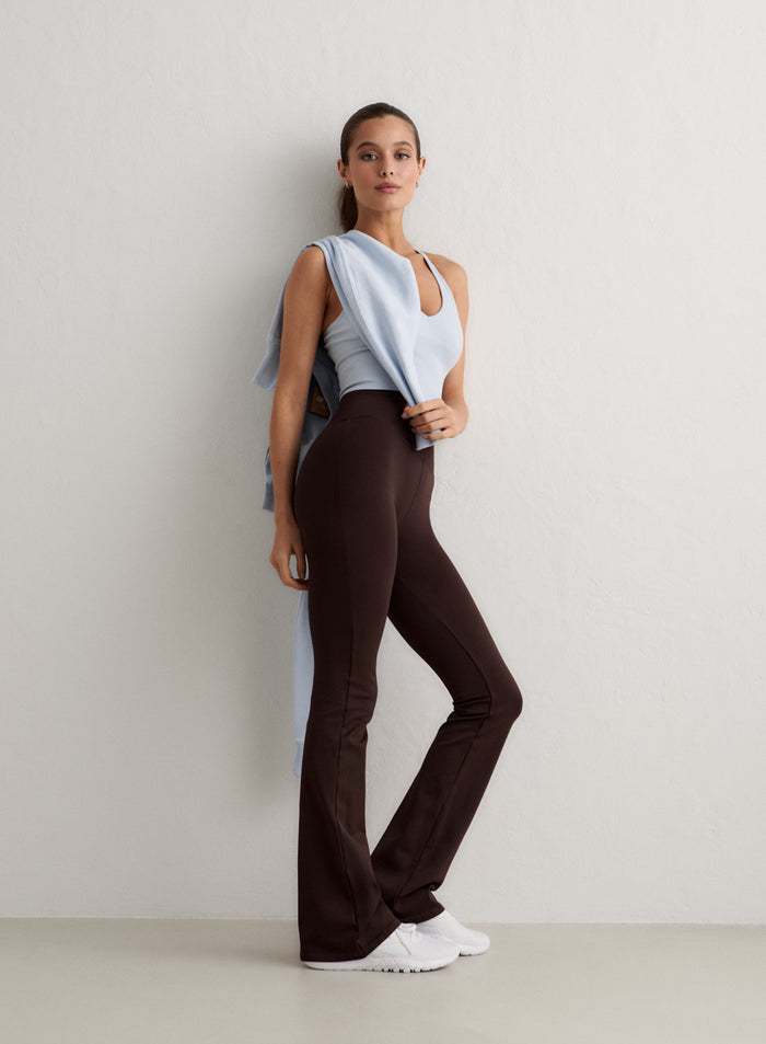 Nainen poseeraa valkoista seinää vasten yllään suklaanruskeat aim'n Shape Seamless Flare Tall Tights -trikoot, vaaleansininen toppi ja valkoiset tennarit [S-174cm]