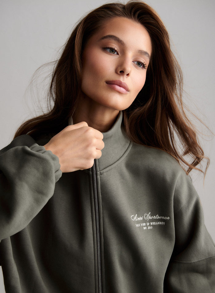 Lähikuva naisesta, jolla on oliivinvihreä korkeakauluksinen aim'n Script Zip Jacket logolla. [S-174cm]