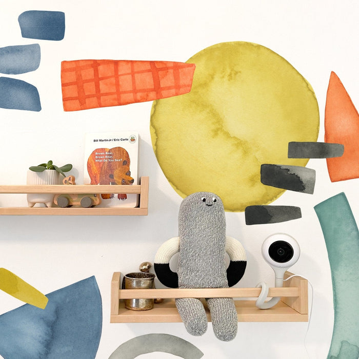 Modern nursery wall decor with colorful watercolor decals, light wood floating shelves displaying a knitted gray plush toy, Brown Bear board book, small potted plant and white baby monitor camera