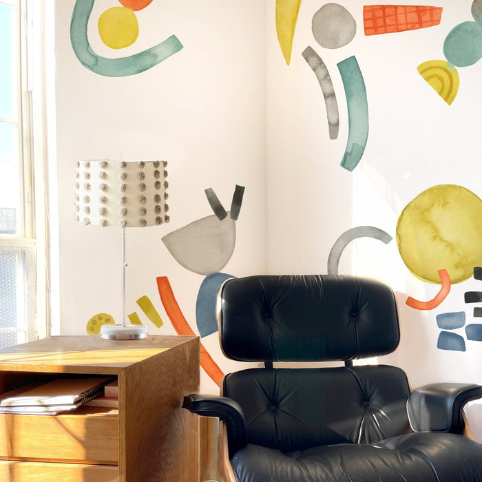 Black leather mid-century lounge chair next to a wooden side table with textured white lamp, colorful abstract watercolor wall mural and sunlight in a minimalist home office