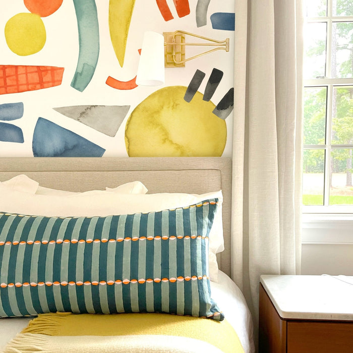 Modern bedroom with colorful watercolor mural accent wall, beige upholstered headboard, teal striped lumbar pillow, cream curtains and wooden bedside table by window