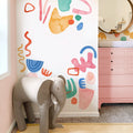 Modern baby nursery corner with gray elephant-shaped chair, colorful abstract wall decals and pink dresser with changing pad