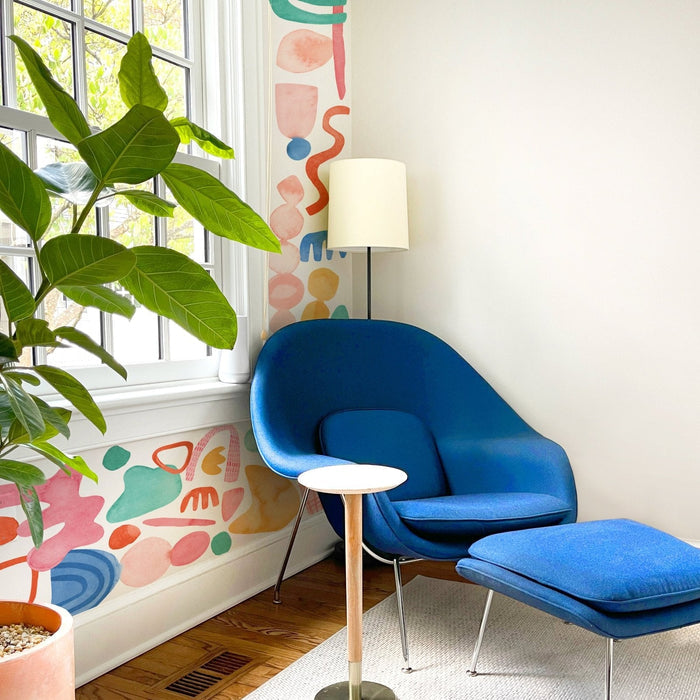 Cozy modern reading nook with blue lounge chair and ottoman, colorful abstract wall mural, floor lamp and large green houseplant by sunlit window