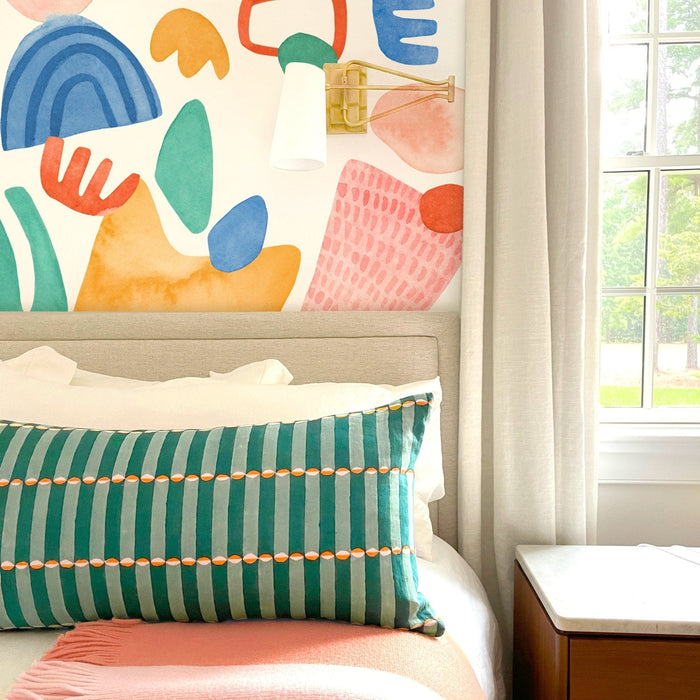 Colorful abstract watercolor mural bedroom with beige upholstered headboard, green striped lumbar pillow, pink throw blanket, brass swing wall sconce and sunlit window