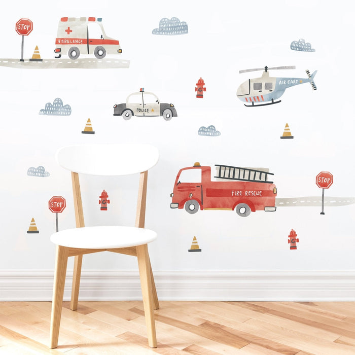 Kids room nursery wall decal in watercolor style featuring emergency vehicles — red fire rescue truck, police car, ambulance and air care helicopter with stop signs and traffic cones on a white wall behind a wooden chair