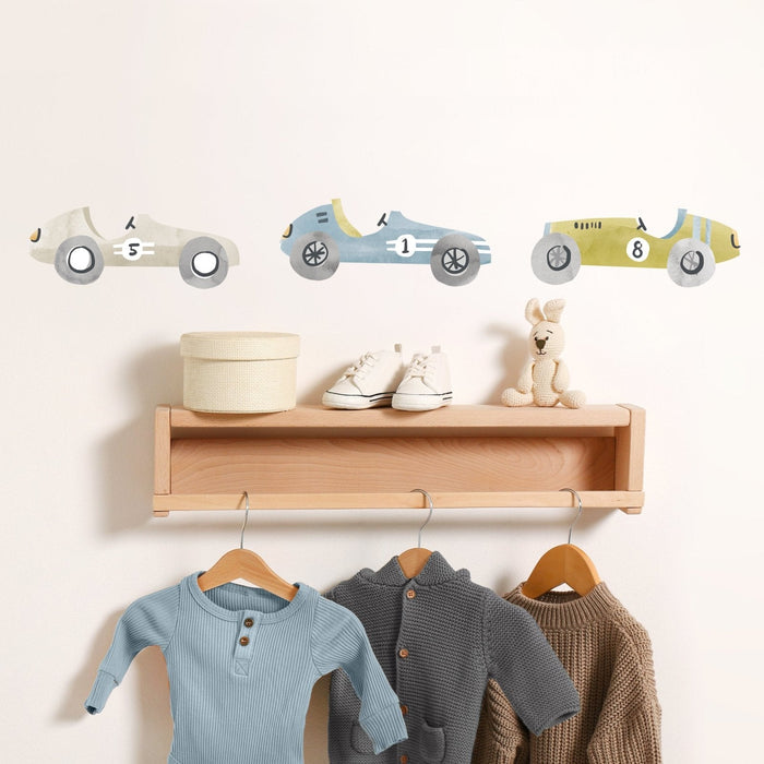 Neutral nursery decor with watercolor race car wall decals above wooden shelf holding baby shoes, round storage box, plush bunny and hanging baby clothes