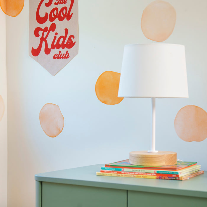 Kids room decor: white table lamp on a mint-green dresser with stacked children's books, orange watercolor circle wall decals and a "The Cool Kids Club" fabric banner