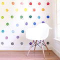 White modern Eames-style chair with wooden legs on light hardwood floor beside window, colorful watercolor polka dot wall decals in minimalist nursery