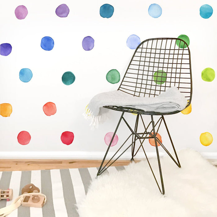 Modern nursery playroom featuring colorful watercolor polka dot wall decals, black wire mid-century chair draped with a gray throw, gray-and-white striped rug, white faux fur rug and wooden toys