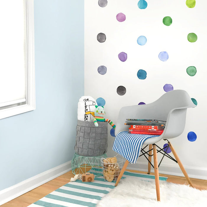 Nursery reading nook with white Eames-style chair, blue-and-white striped rug and blanket, colorful watercolor polka dot wall decals, gray felt toy storage basket, wooden floor and natural light