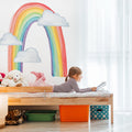 Child reading on bed in bright kids bedroom with rainbow wall mural, stuffed animals, and colorful storage