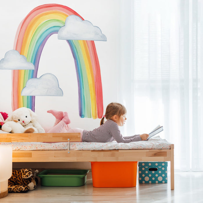 Child reading on bed in bright kids bedroom with rainbow wall mural, stuffed animals, and colorful storage
