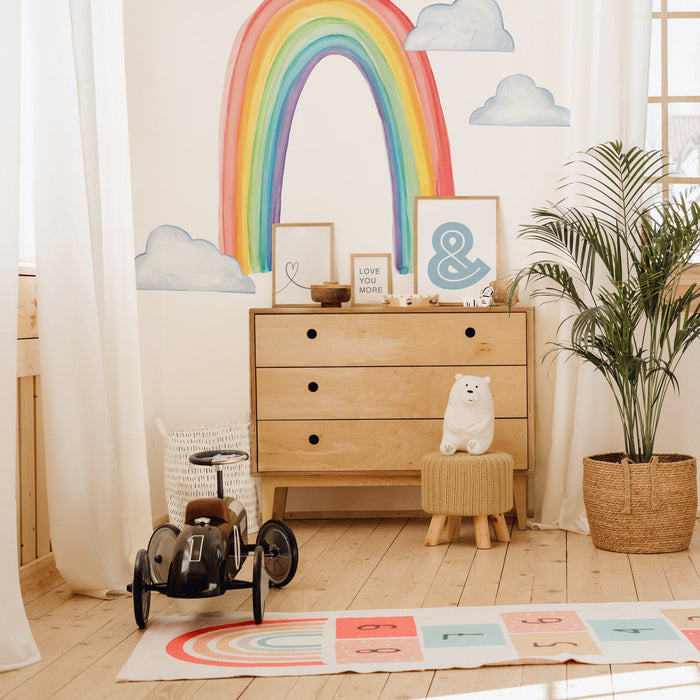 Bright children's nursery with rainbow wall mural, wooden three-drawer dresser, vintage toy car, plush bear, framed prints, and potted palm plant