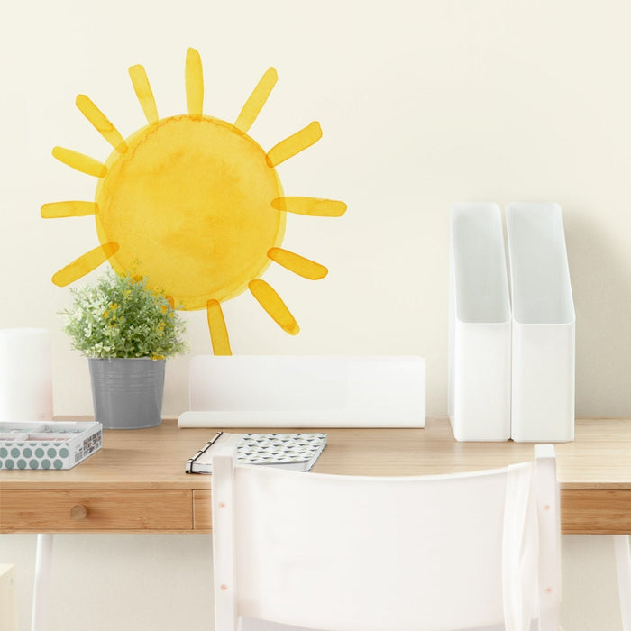Home office desk with yellow sun wall decal, gray potted plant, white desk organizers and notebook on light wood surface
