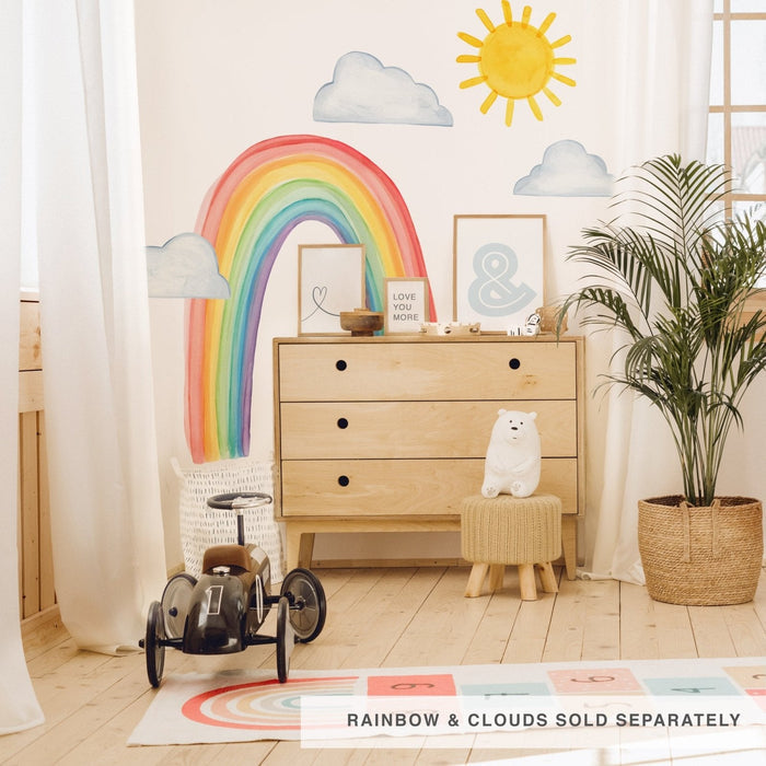 Bright gender-neutral nursery decor featuring a rainbow wall decal and sun-and-cloud mural above a light wood dresser with framed prints, black ride-on toy car, plush bear and potted palm plant
