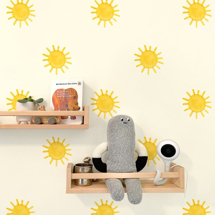 Nursery decor with yellow sun wall decals, light wood floating shelves holding a potted plant, wooden toy car, "Brown Bear" board book, knitted gray doll and white baby monitor.