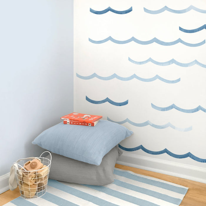 Coastal nursery corner with blue wave wall decals, striped rug, stacked pillows, wire basket of wooden toys and children’s books
