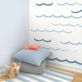 Coastal nursery corner with blue wave wall decals, striped rug, stacked pillows, wire basket of wooden toys and children’s books