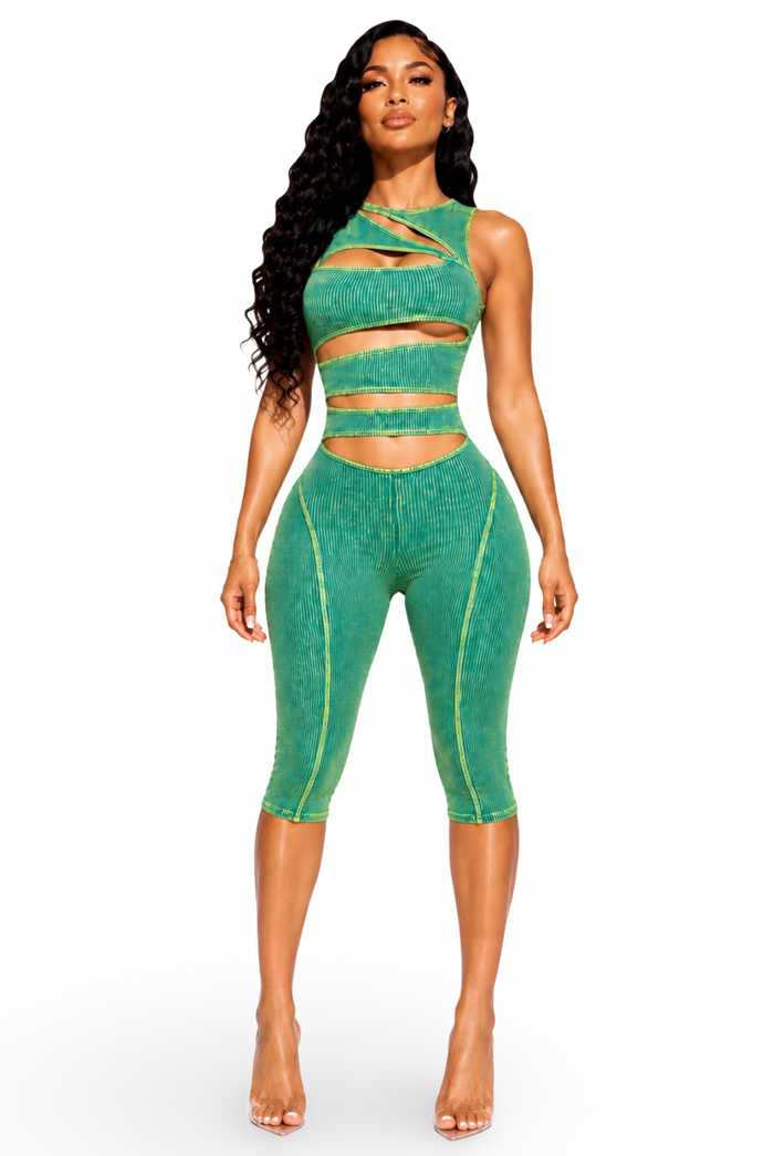 A woman modeling a matching green two-piece activewear set consisting of a high-waisted crop top with cutouts and strappy details, and matching high-waisted capri leggings with side seam details, shot against a plain white background.