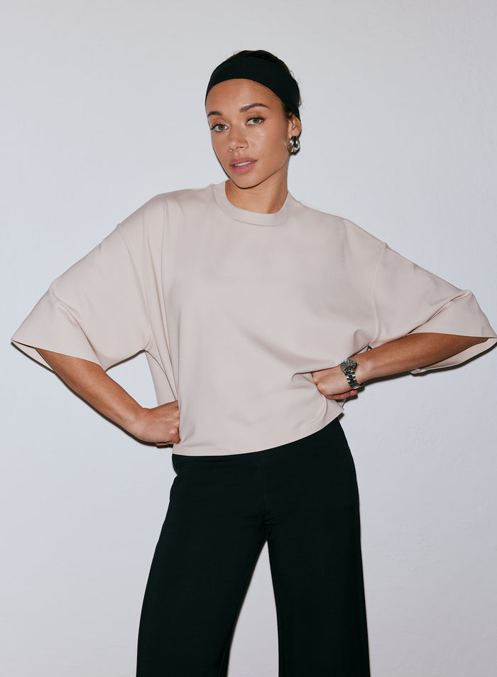 Nainen yllään hiekanvärinen Luxe Boxy Short Sleeve -toppi ja mustat housut, kädet lanteilla [M-170cm]