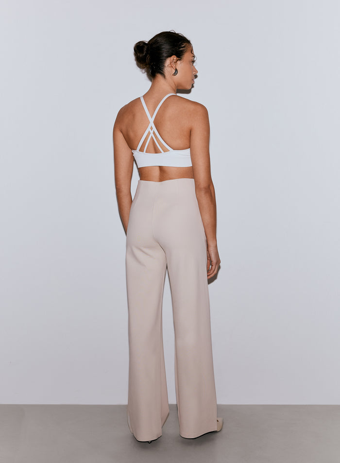 Nainen takaapäin yllään hiekanväriset, leveälahkeiset Luxe Pants -housut ja valkoiset monihihnaiset urheiluliivit. [36-170CM]