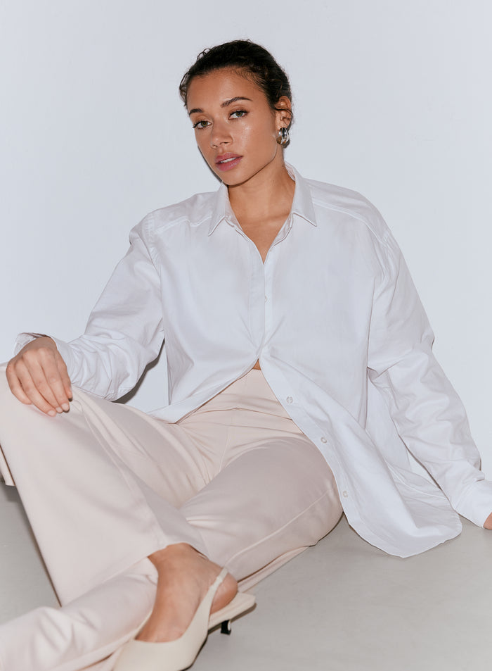 Nainen, jolla on tummat hiukset, yllään valkoinen oversized-paita ja hiekanväriset Luxe Pants -housut, istuu lattialla. [36-170cm]