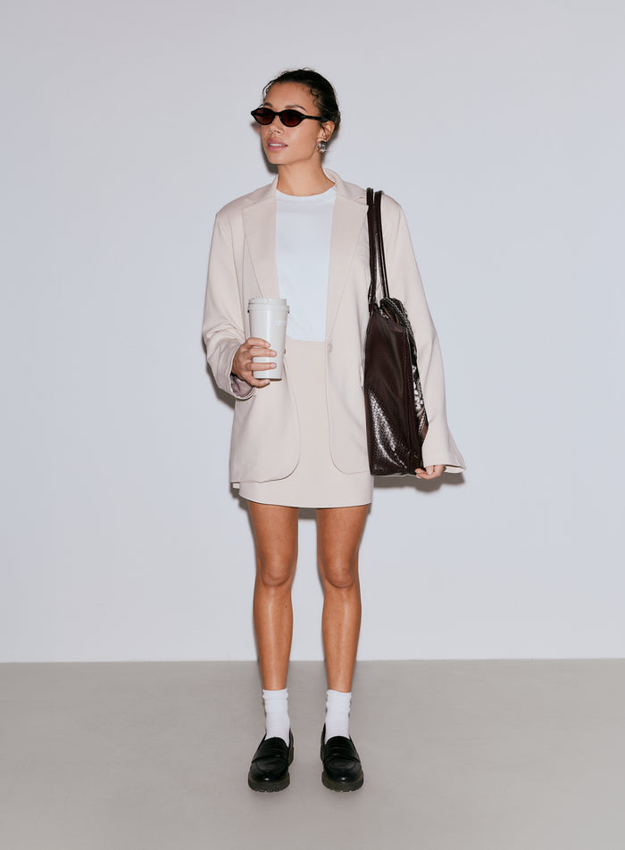 Nainen yllään hiekanvärinen Luxe Relaxed Blazer, valkoinen t-paita ja yhteensopiva hame, pitäen kädessään uudelleenkäytettävää kahvikuppia ja tummanruskeaa kangaskassia. [40-170cm]