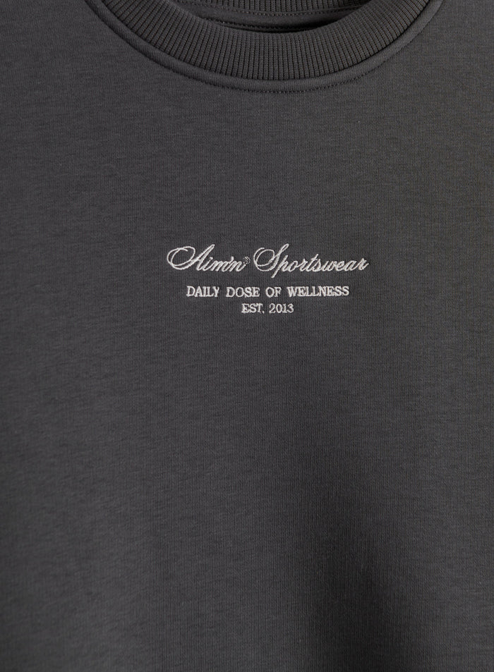 Tummanharmaa Script Sweatshirt, jossa on valkoinen brodeerattu aim'n-logo ja joustinneulottu pyöreä pääntie.