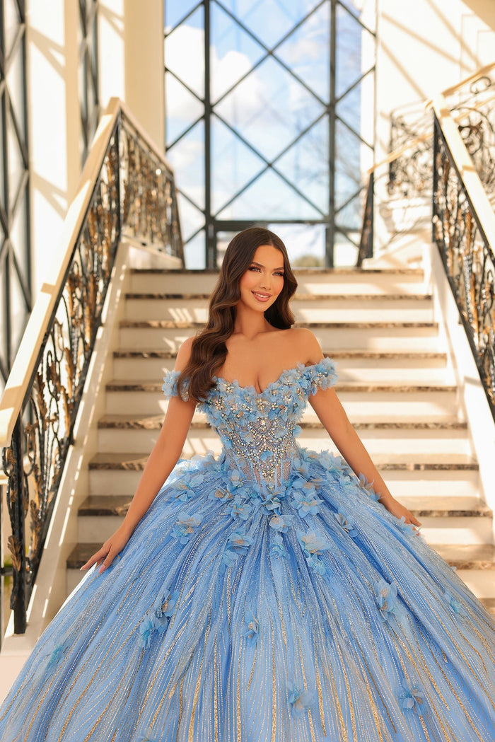 A woman in a voluminous blue floral quinceañera gown stands on an elegant staircase with ornate railings.