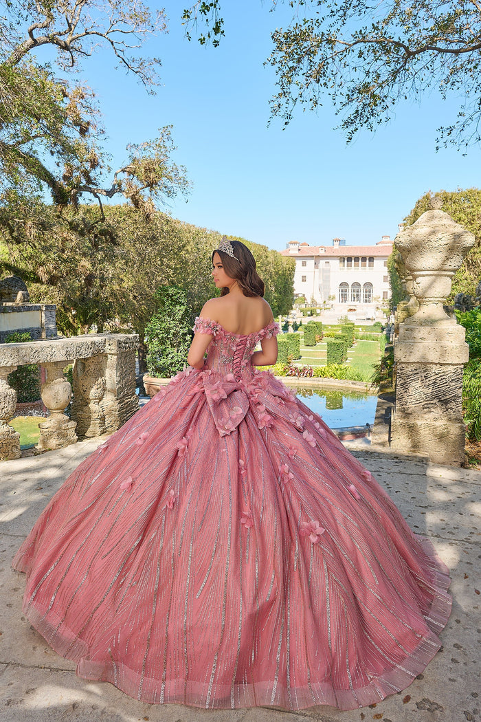 A person in a voluminous dusty rose quinceañera gown stands in a formal garden with a grand white mansion behind.