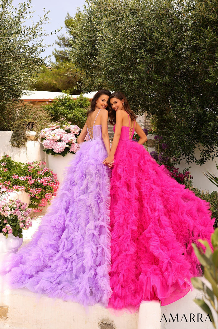 Two women in lavish ruffled gowns, one purple and one pink, stand close together in a garden of flowers.