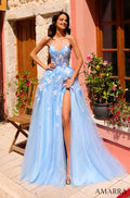 A woman wears a light blue floral lace gown with a high slit, standing in a sunlit courtyard with flowers.